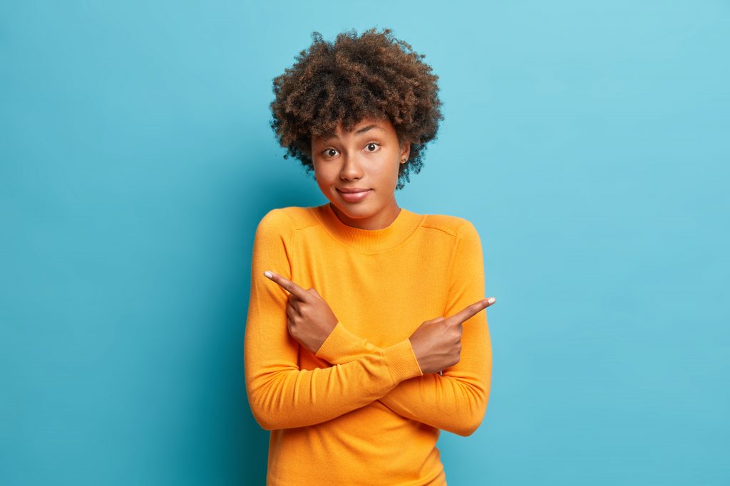 rapariga morena com cabelo encaracolado com camisola amarela hesitante apontando para os dois lados com ar de dúvida