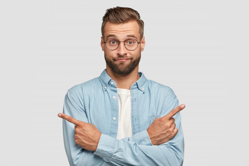 rapaz louro com oculos e camisa azul hesitante apontando para os dois lados com ar de dúvida